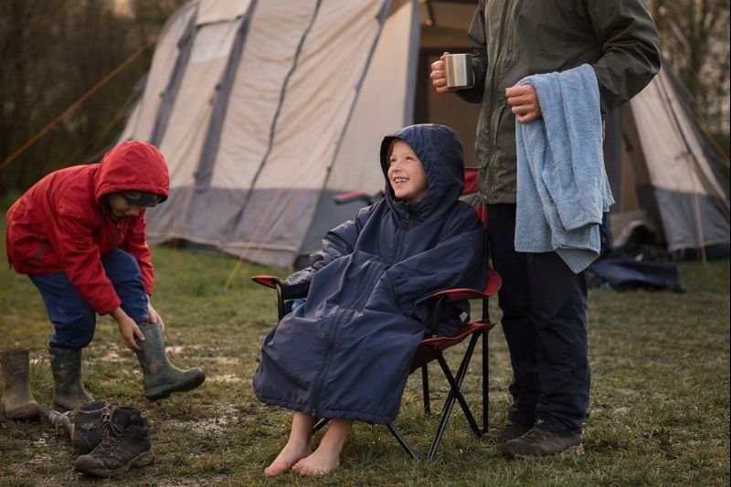 Family camping scene with a child sitting in an outdoor changing robe while others remove muddy boots near a tent