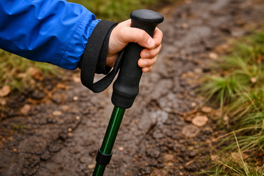 Close up of a child's hand correctly gripping an EVA foam hiking pole handle with wrist strap on a muddy outdoor trail