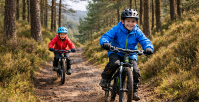 Two children wearing helmets and waterproof jackets riding mountain bikes on a muddy woodland trail through pine trees in Scotland
