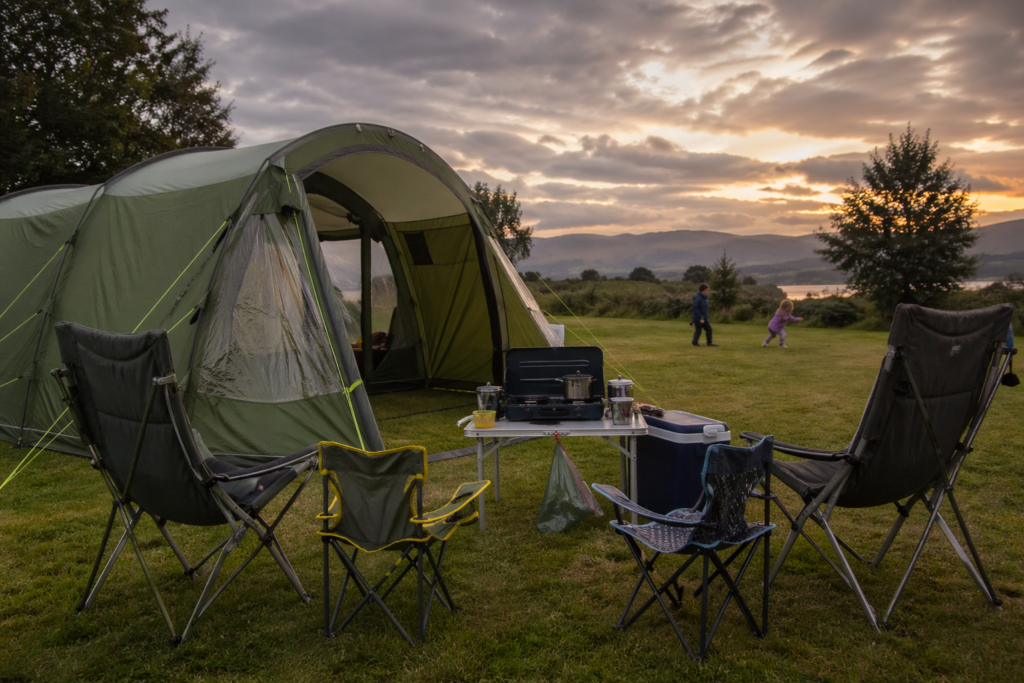 UK family campsite setup at dusk with green tent camping chairs folding table cool box and two children playing in background