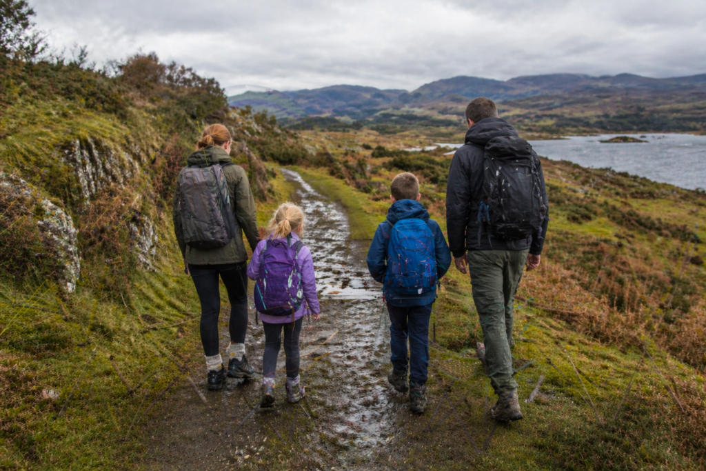 Family with two children wearing walking boots and backpacks on a muddy lochside countryside path in overcast Scottish daylight