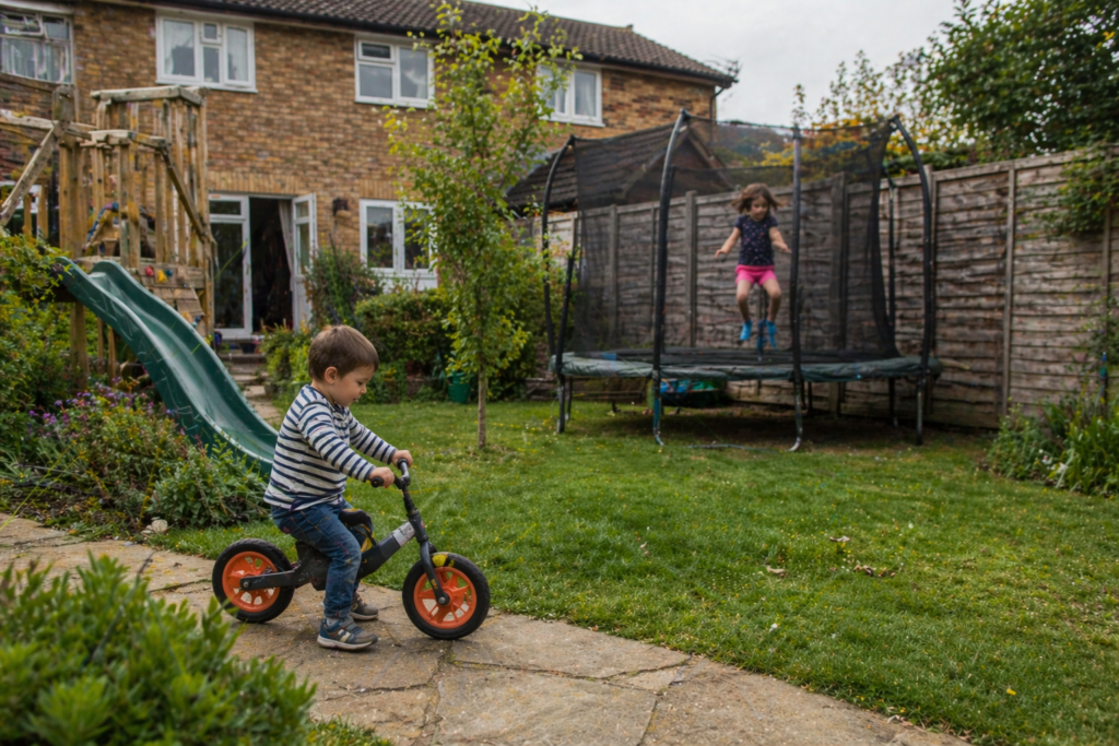 Two children playing in a UK family garden one on a balance bike and one jumping on a trampoline with climbing frame visible in background