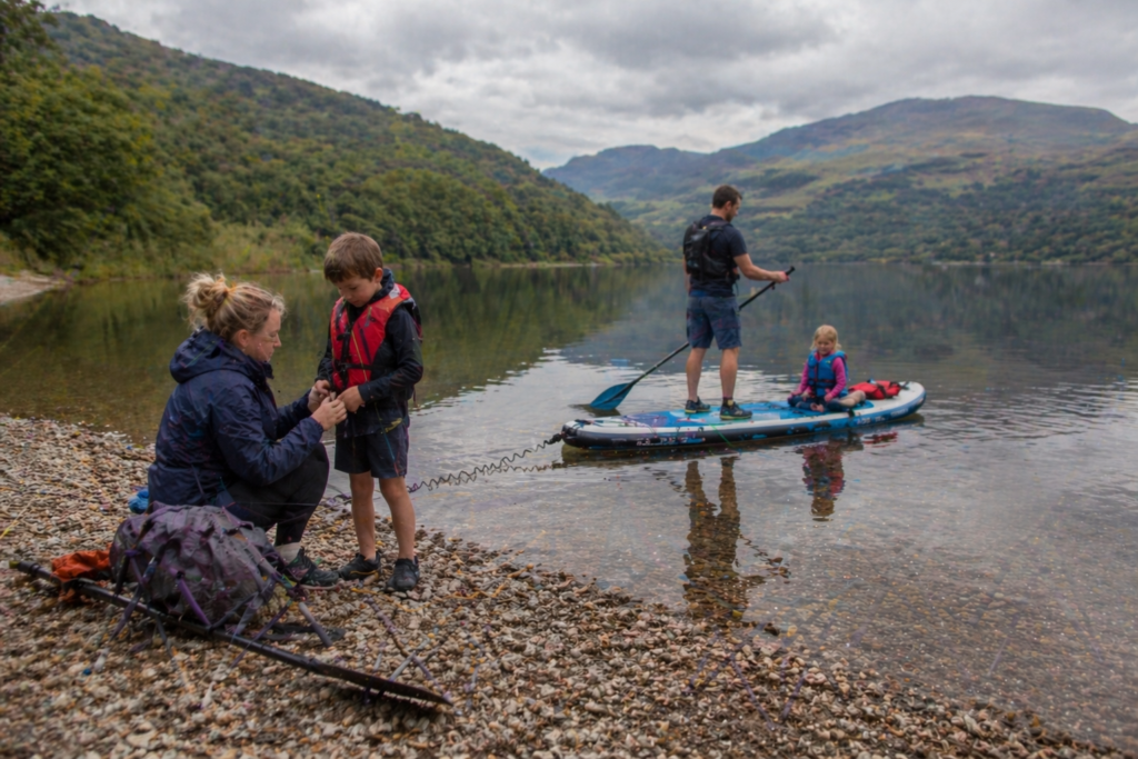 Family on a calm Scottish loch with adult and child on inflatable paddle board and family visible on shore with hills reflected in still water
